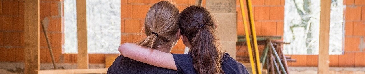 Zwei befreundete Frauen umarmen sich gegenseitig in Gebäude vor orangener Wand